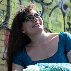 Woman in blue shirt, leaning against a colorful graffiti wall, smiling up at the sun.