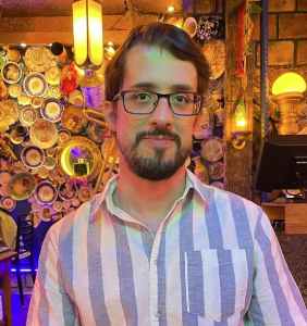Man in striped shirt, with short dark hair, standing in front of a backdrop of yellow and blue.