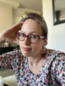 A woman with brown hair, pulled back from her face. She is wearing stylish raspberry-colored glasses and a multicolored shirt.