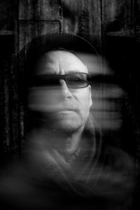 Black and white photo of a man wearing sunglasses. The image is blurred, with the appearance of motion.