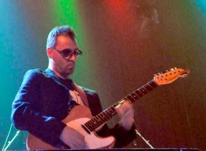 A man wearing sunglasses, playing a cool guitar. Green and red stage lights fill the background.