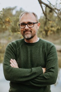A man with a green sweater, dark-rimmed glasses, standing in the foreground. Trees and brush are behind him in the distance.