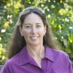 A white woman with long  dark hair, smiling. The background is slightly-blurred green tree leaves. L.D. is wearing a purple collared shirt and earrings.