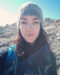 A woman with long dark hair and light skin, wearing a knit cap and backpack. Blue sky and a rocky slope are behind her.