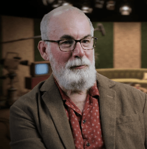 A man with short grey hair and beard, wearing glasses and a rust-colored buttoned shirt under a lovely brown jacket. He looks like a nice guy!