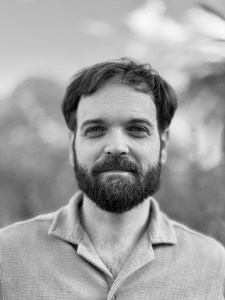 A black & white photo of a happy-looking guy with dark short hair and beard, wearing a collared shirt. The background is blurry mountains.