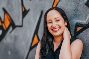 A smiling Latine woman with long brown hair, wearing a black sleeveless top, stands in front of a gray, orange, and black graffiti art background.