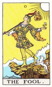 A tarot card with the words “The Fool” at the bottom of the image. The illustration is a person in medieval costume, holding a white flower and carrying a travel back on a stick. The person is looking up, into a sunny sky, but they are approaching the edge of a cliff, giving the impression they might fall with their next step. I white dog plays near the person’s feet.