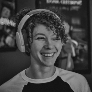 A black and white image of a young woman with short curly hair and a beautiful smile. She was probably laughing when the photo was taken, inviting everyone to laugh with her. She is wearing a headset, which is pretty cool.
