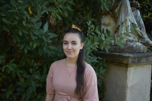 A photo of a woman with long dark hair over her left shoulder. She wears a pink shirt, and stands in front of greenery and garden sculpture. You would probably love her writing and want to hang out with her.