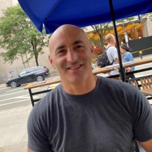 A photo of a smiling man in a gray shirt. He is positioned in front of a city street with a wooden bench and blue awning. He looks happy!