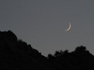 A slim, pale-gold crescent moon hangs in a gray twilight sky above the black silhouette of desert hills. Taken at Dinosaur National Monument in Utah.