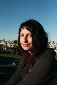 Headshot of T. K. Rex on a sunny day at a park with the San Francisco skyline in the background. The author has long brown hair and is surprisingly pale considering how bright that sun looks, as if they have not in fact been to the park in some time, but according to the smile they are glad to be there nonetheless. They have brown eyes, a rather protruding nose and a grey hoodie. Credit: Giovanna Lomanto. 