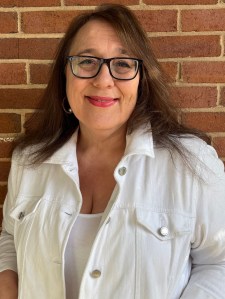 Caroline Bock author photo features a woman with long russet hair wearing a white shirt, standing in front of a red brick wall. She has a beautiful smile.