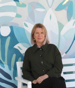 Woman with short blonde hair looking into camera. She is wearing a green jacket. The background is a blue abstract swirl.