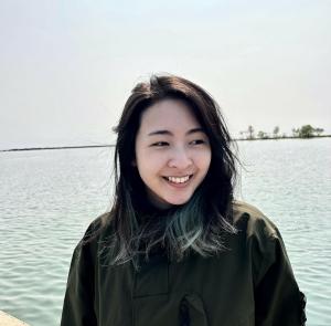 An Asian woman with a beautiful smile, standing in front of a light blue body of water.