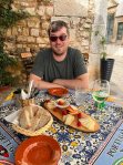Photo of Ben at an outdoor table with various dishes of food. The image is colorful and looks like fun! Ben is wearing a short sleeved shirt and sunglasses.