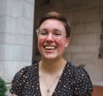 Headshot of Kay, a white person with short brown hair, brown eyes, and rosy cheeks. They’re wearing round clear-framed glasses, a silver nose ring, a silver necklace, and a floral dress with puffed sleeves, and they’re laughing.