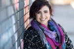 Headshot of Naseem, a light-skinned West Asian person with curly brown hair, brown eyes, and a nose stud. They’re wearing a textured black sweater, a colorful scarf, and tombstone earrings, and smiling widely.