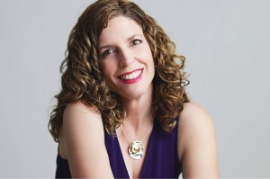 A white female-presenting person with long curly reddish-brown hair. She is smiling. She is wearing a purple shirt and necklace. 